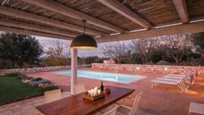 An outdoor patio area with a wooden table, white chairs, a stone wall, and a pool. - TD Lamiamola Stylish Trulli & Lamie With Pool (Ostuni)