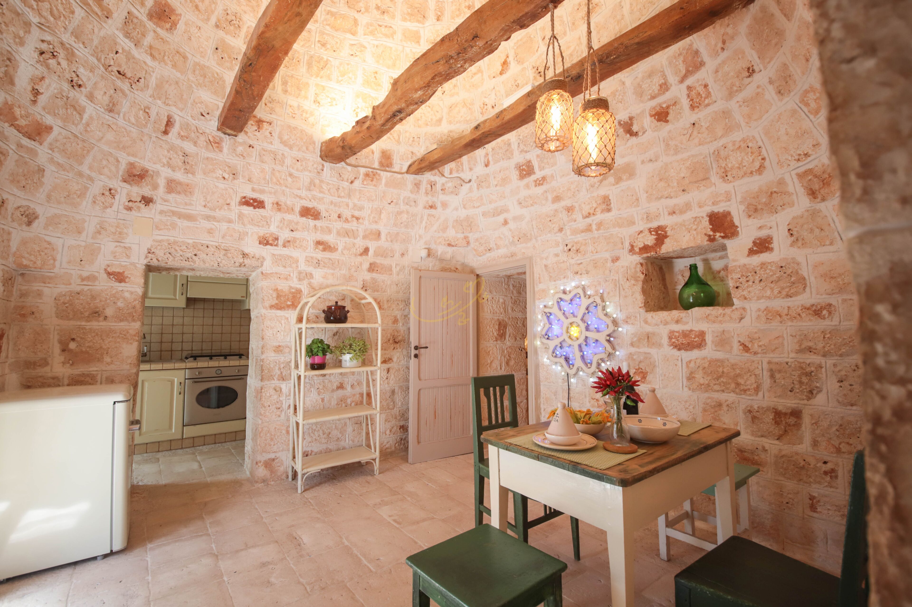 A rustic kitchen with exposed brick walls, wooden beams, a small dining table with chairs, and a hanging light fixture.