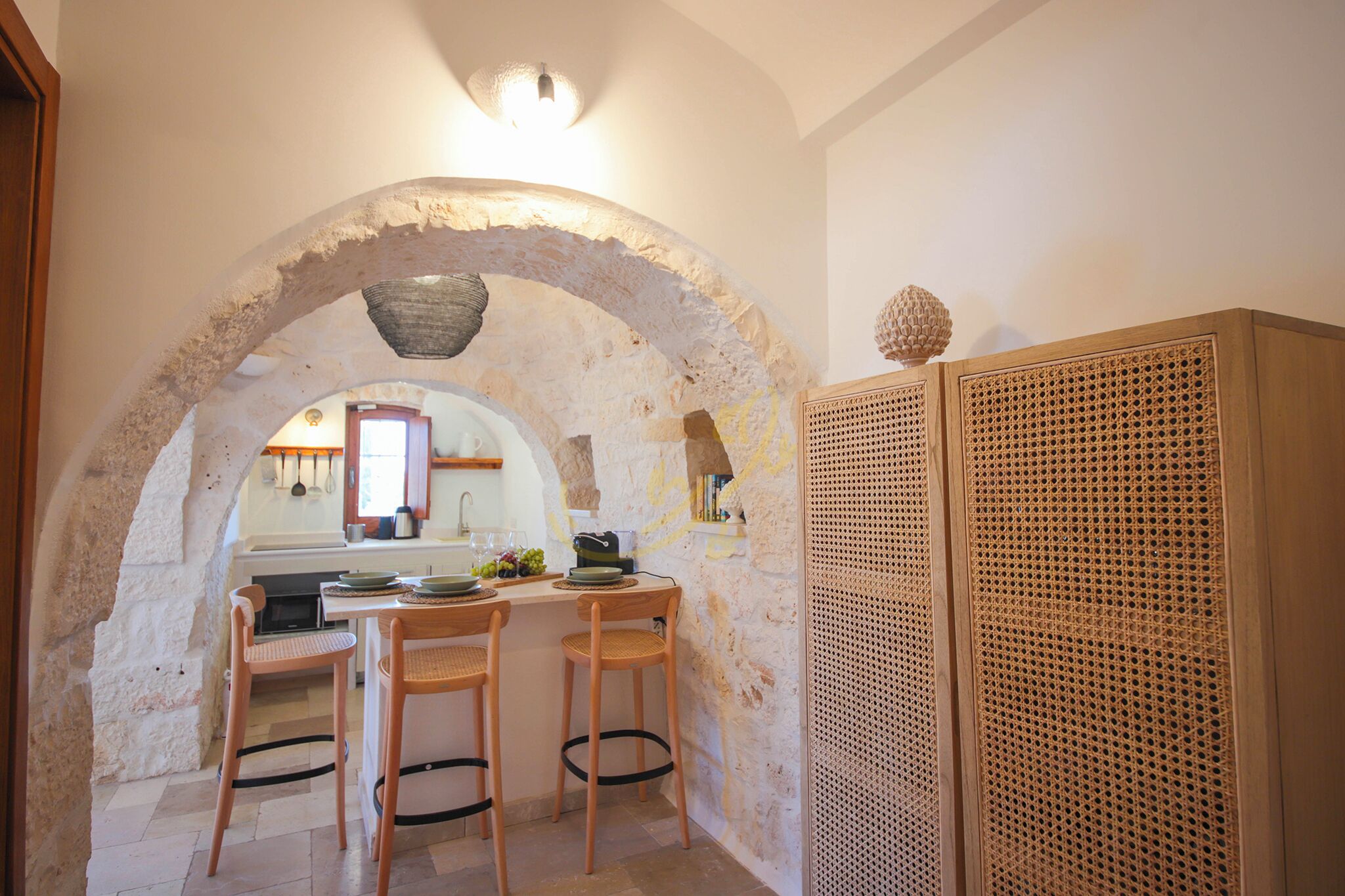 A kitchen with an arched stone ceiling, wooden chairs, a table with plates and glasses, and a woven basket on the counter.