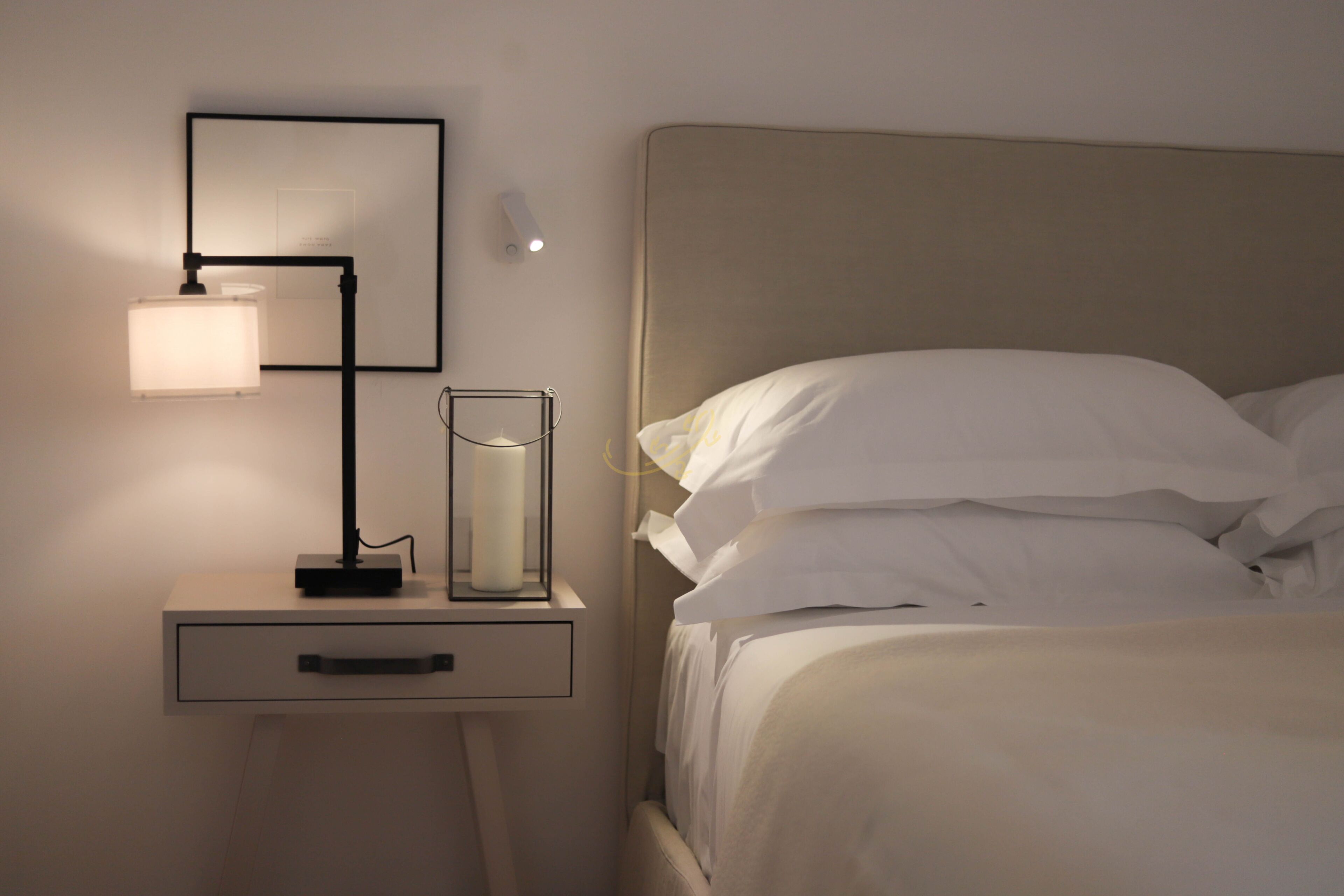A neatly made bed with white linens, a bedside lamp, a framed picture, and a small bedside table with a drawer.