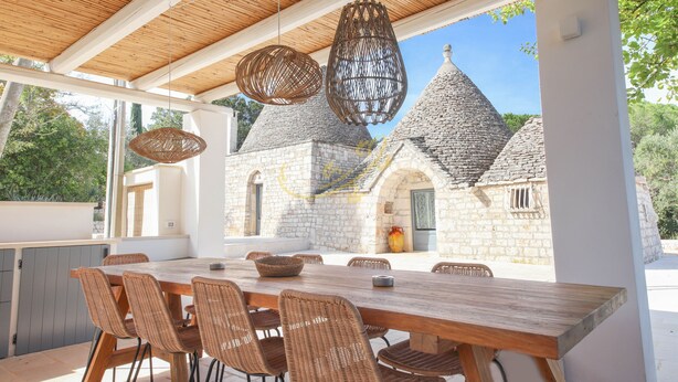 An outdoor dining area with a wooden table and wicker chairs, overlooking a stone building with conical roofs. - TD Trulli Tortorella With Nature Pool & Sauna (Monopoli)