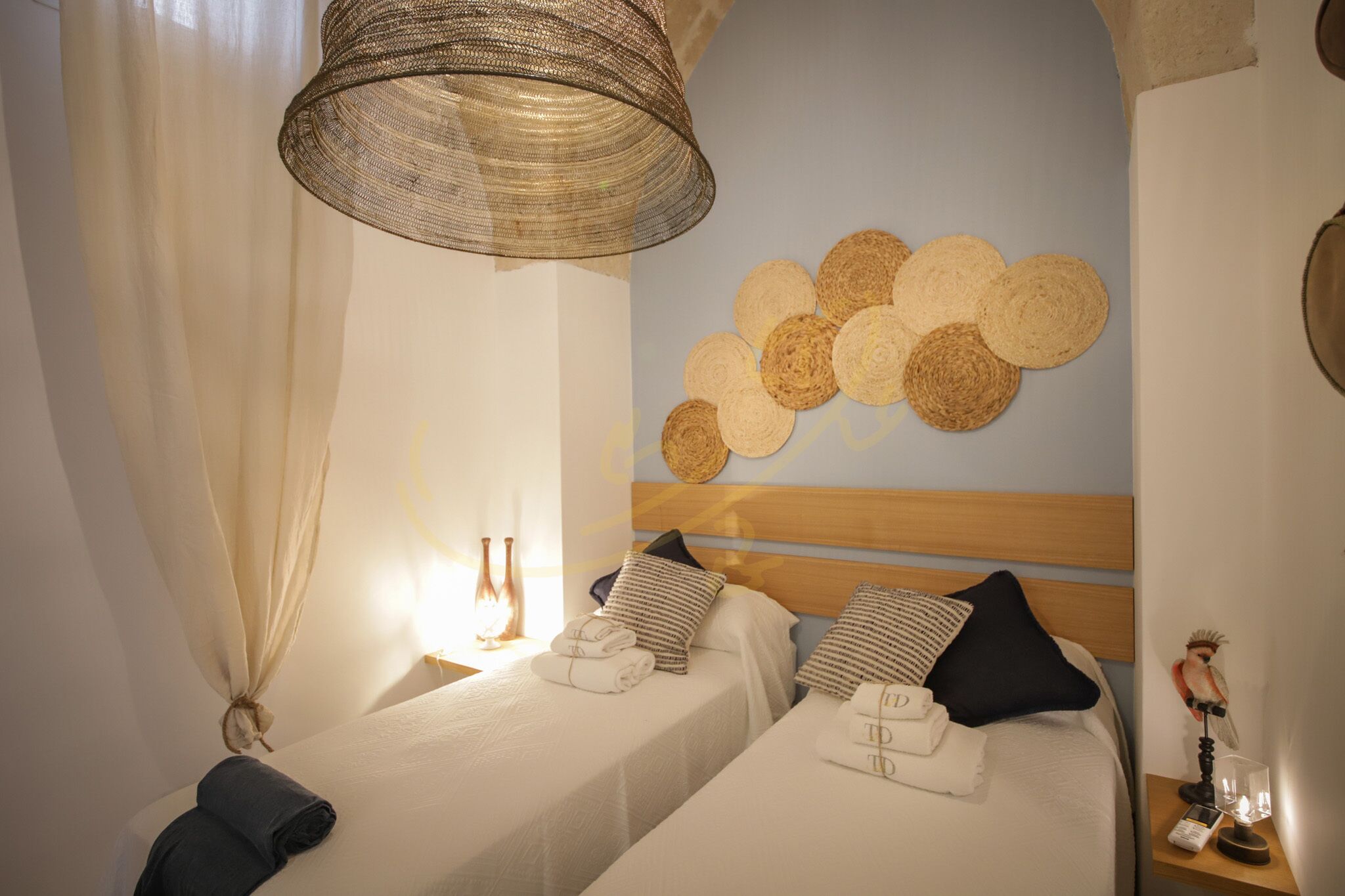 A hotel room with two beds, a woven headboard, a wall-mounted woven decoration, and a nightstand with a lamp and a figurine.