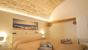 Un dormitorio con techo de piedra, un cuadro con escena de playa, una cama con ropa de cama color crema y toallas con un logotipo.