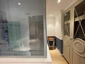 Ein Zimmer mit großem Fenster, Jalousien, einem Holzschrank mit Spiegel und einem kleinen Tisch.