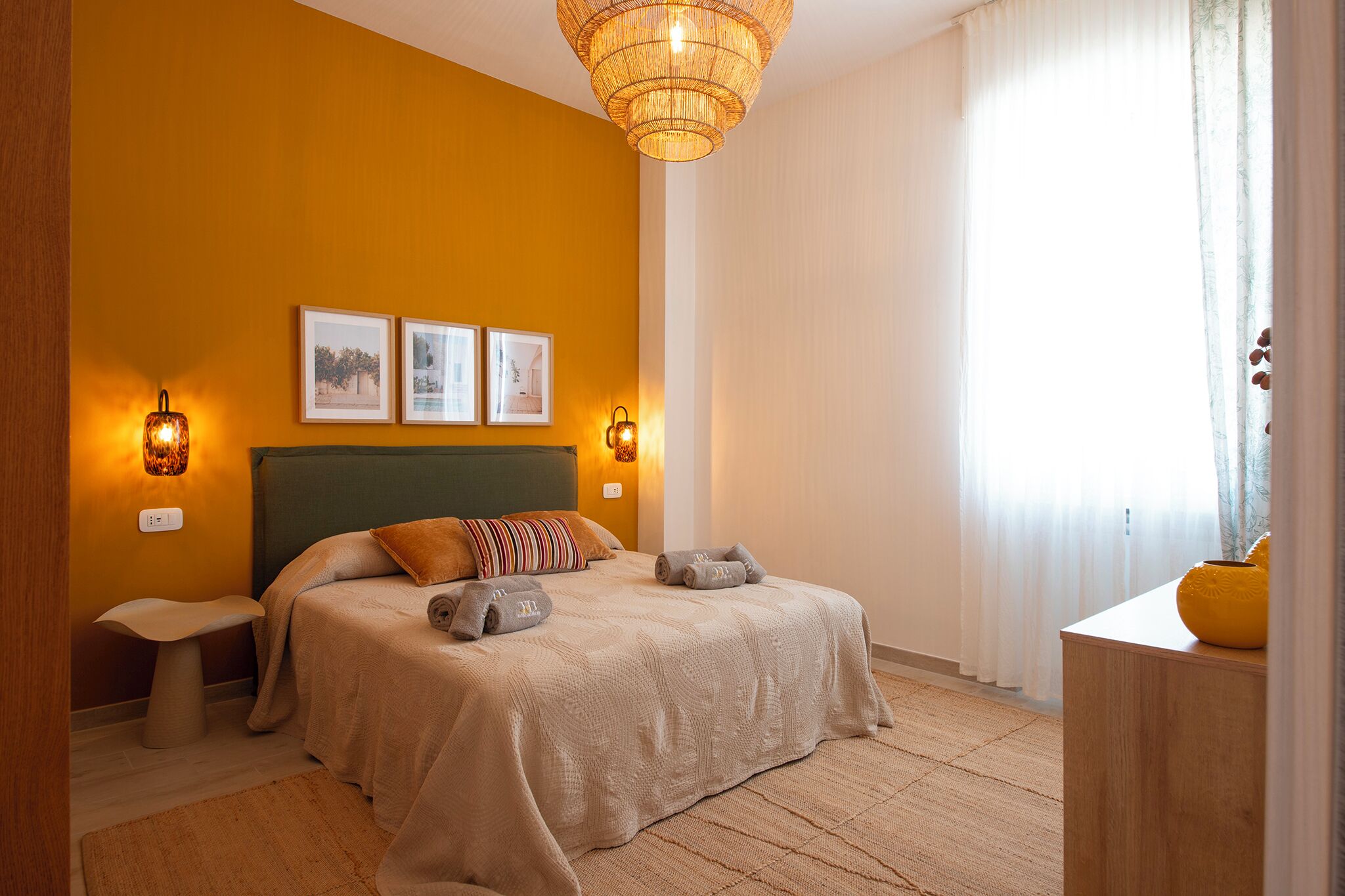 Un dormitorio con cama, mesitas de noche, lámpara colgante y cuadros en la pared.