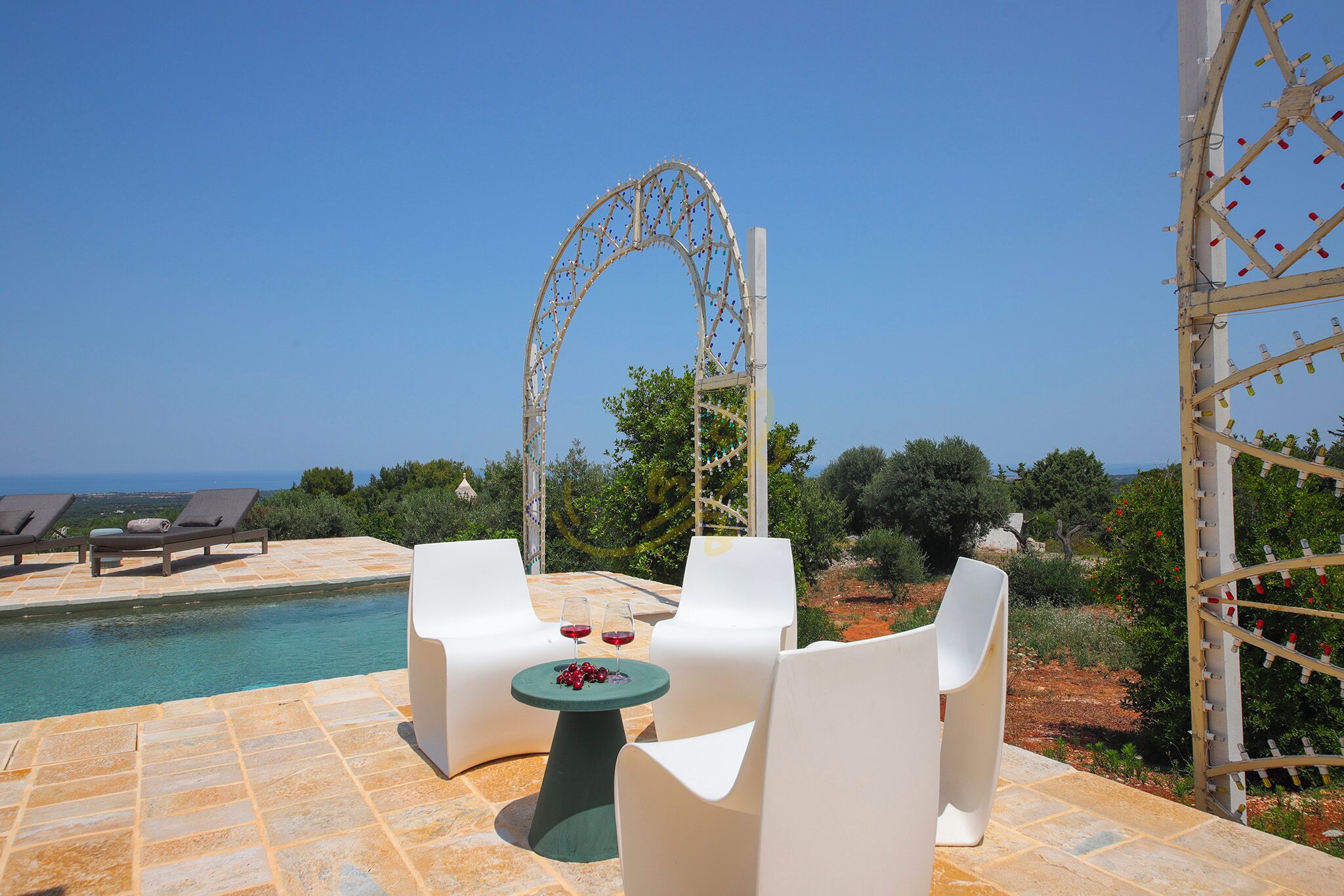 A poolside area with white lounge chairs, a table with two glasses of red wine, and a decorative archway.