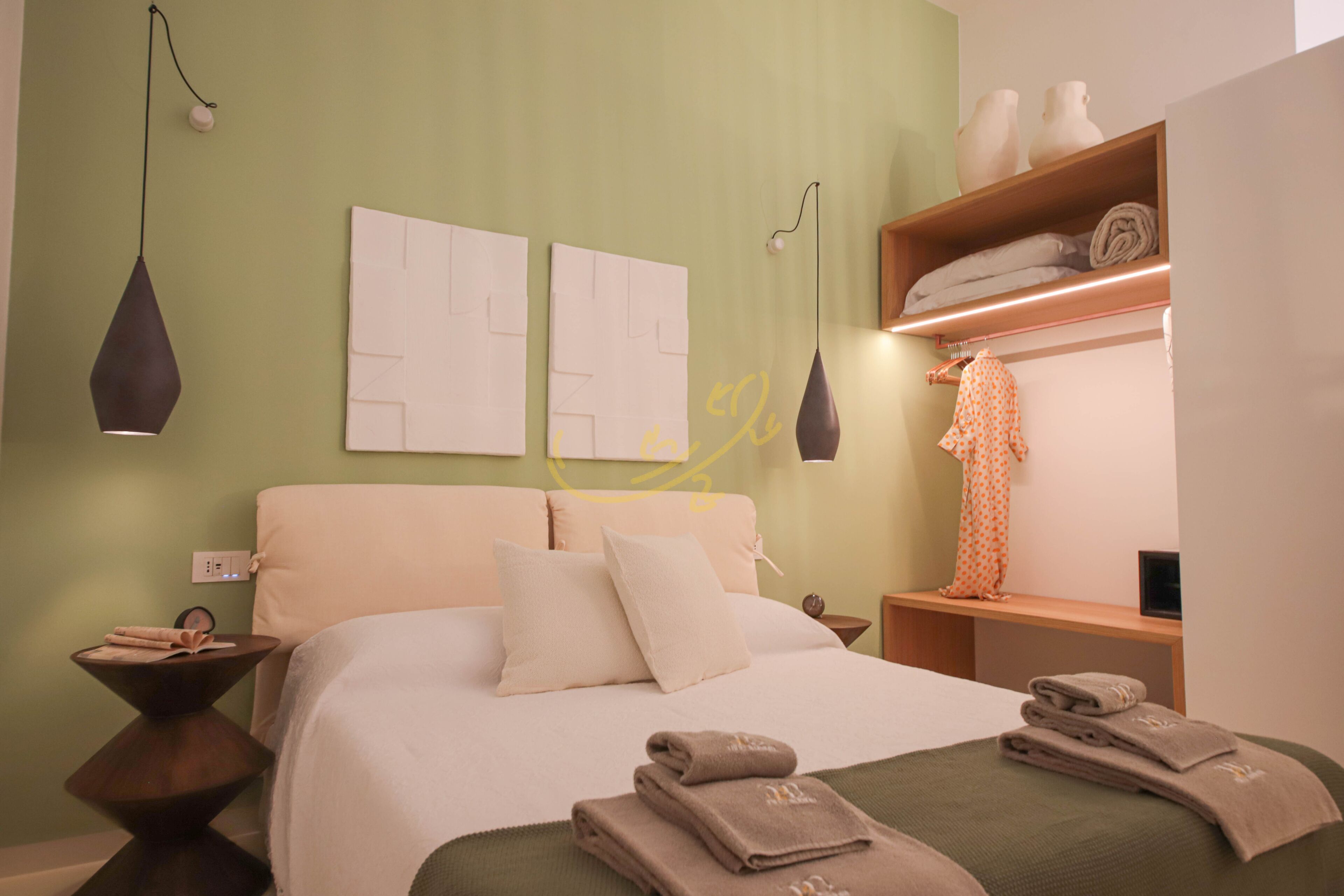 Une chambre d’hôtel comprenant un lit, des tables de chevet, une commode, une étagère avec des serviettes et des œuvres d’art murales.