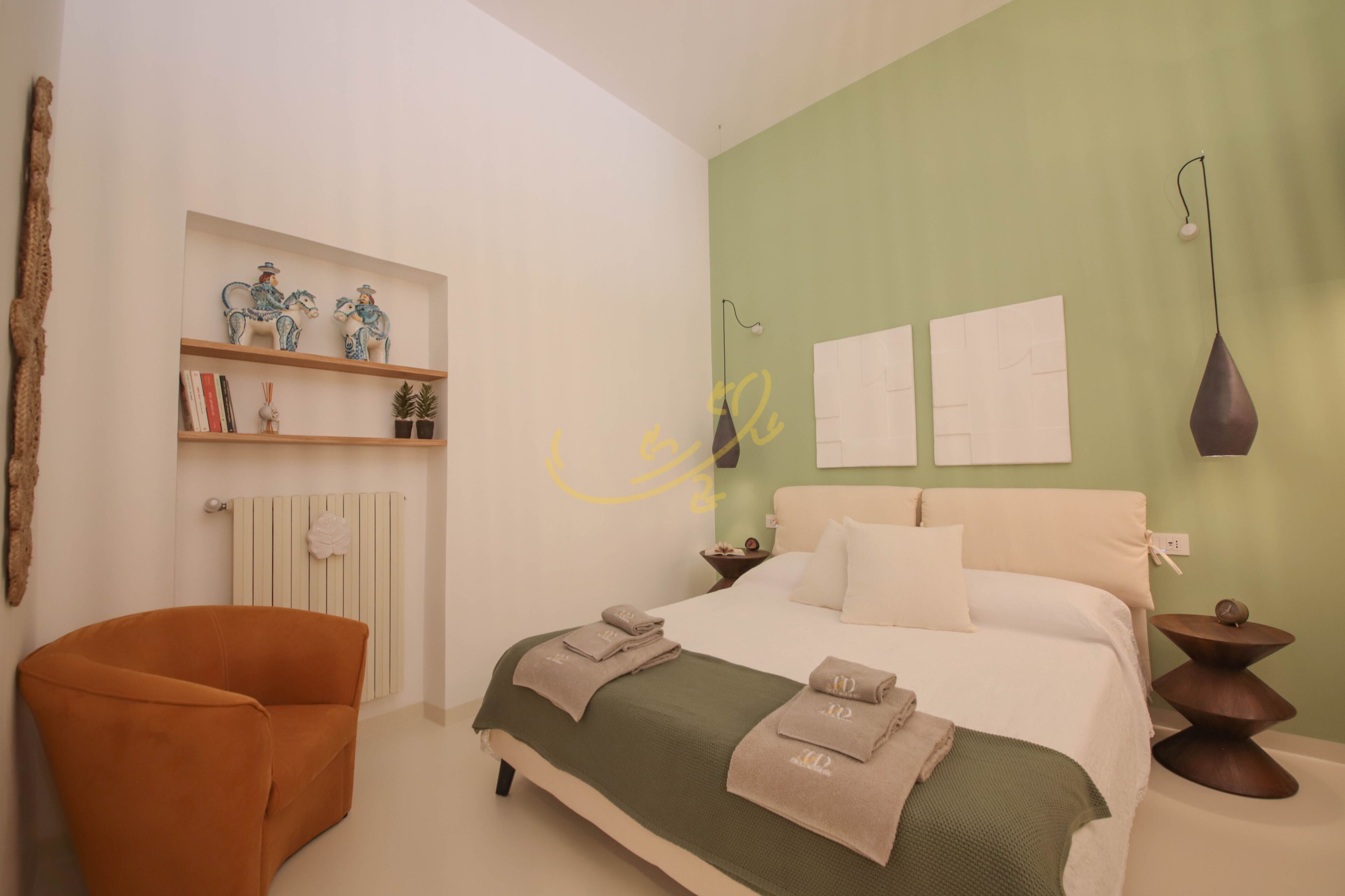 Un dormitorio con una cama, una silla, una estantería con objetos decorativos y una pared con dos cuadros enmarcados.