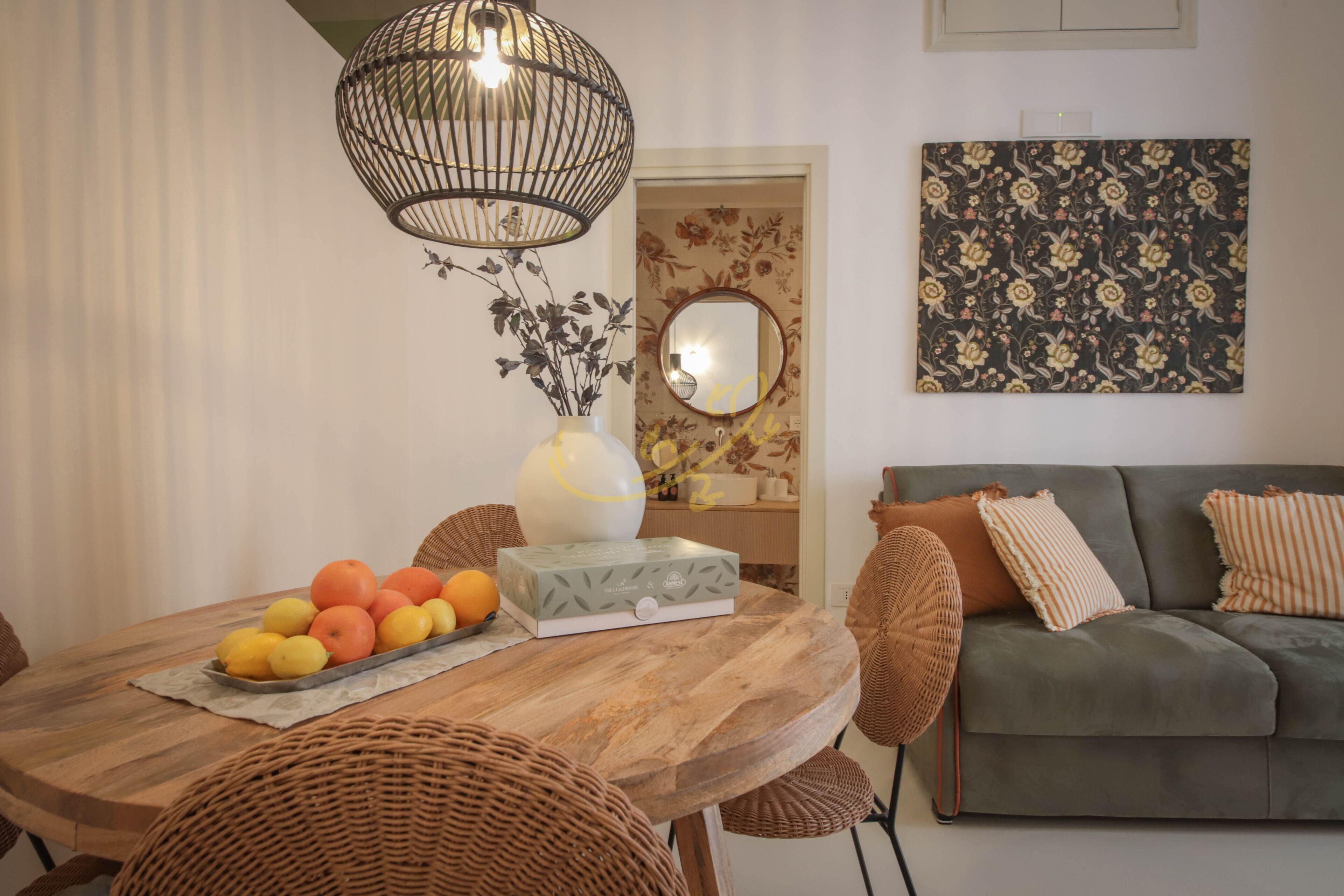 A dining area with a wooden table, wicker chairs, a fruit bowl, and a vase with flowers.