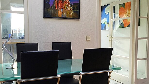 Uma sala de jantar com mesa de vidro e cadeiras pretas, um quadro de um edifício e uma porta com painéis de vidro.