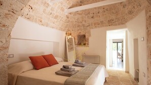Un dormitorio con pared de piedra, cama con cubrecama color beige, almohada roja y espejo con lámpara.