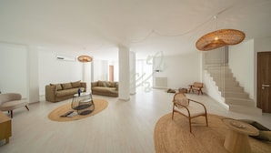 Un vaste espace de vie comprenant un canapé, une table basse et des fauteuils, un escalier et un grand luminaire tressé.