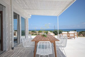 Een overdekt terras met een houten tafel en witte stoelen, met uitzicht op een prachtig zeegezicht en landschap.