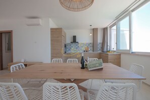 Un espace repas moderne avec une table en bois et des chaises en osier blanches, une cuisine en arrière-plan et de grandes fenêtres.