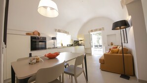 A modern kitchen with white cabinets, a dining table, and chairs. There is a sofa and a lamp in the living area. - TD Trullo Morena Charming Trullo,stunning Sunsets (Monopoli)