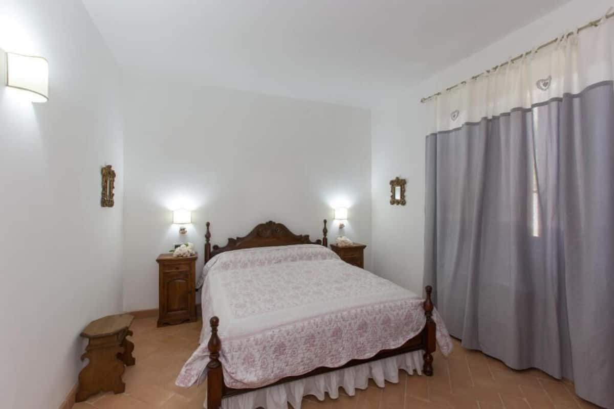 A bedroom with a wooden bed, two bedside tables with lamps, a nightstand, and a window with curtains.