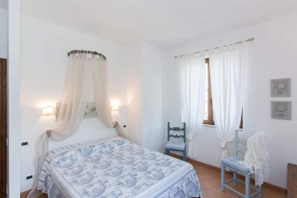 A bedroom with a bed, a chair, a window with curtains, and wall-mounted lights.