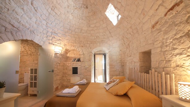 A bedroom with a stone ceiling, a bed with a brown blanket, a nightstand with a lamp, and a view through a doorway. - TD Trulli Della Lama Private Trulli With Pool (Castellana Grotte)