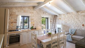 A rustic kitchen with stone walls, wooden beams, a dining table with chairs, a stove, and a white cabinet. - TD Trulli Della Lama Private Trulli With Pool (Castellana Grotte)