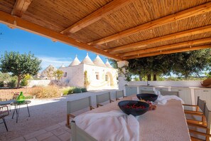 Un área de comedor al aire libre con una pérgola de madera, una mesa dispuesta con un cuenco de fruta y vistas a un edificio blanco con techos cónicos.