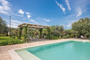 A swimming pool with a pergola and outdoor seating area, surrounded by greenery and trees.