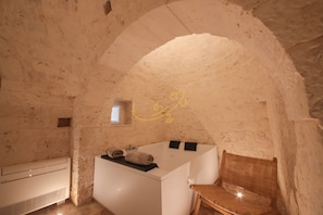 Ein Zimmer mit Steinwänden, einer weißen Badewanne, einem kleinen Fenster und einem Holzstuhl.