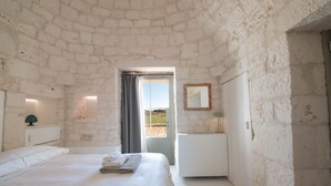 A bedroom with a stone wall, a bed with white linens, a bedside table with a lamp, and a view through a door. - TD I Coni di Pinurè Trulli With Scenic Valley .. (Martina Franca)