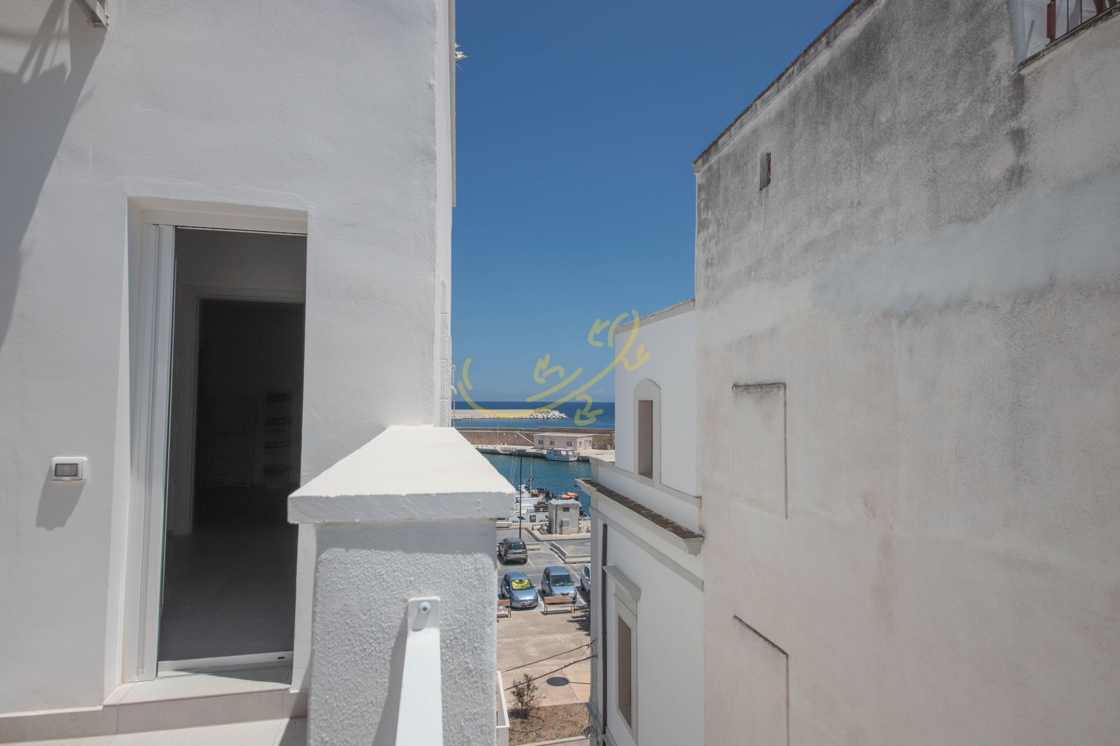 Un callejón estrecho con un edificio blanco a la izquierda y uno gris a la derecha. Se ve un cielo azul despejado arriba, y una masa de agua al fondo.