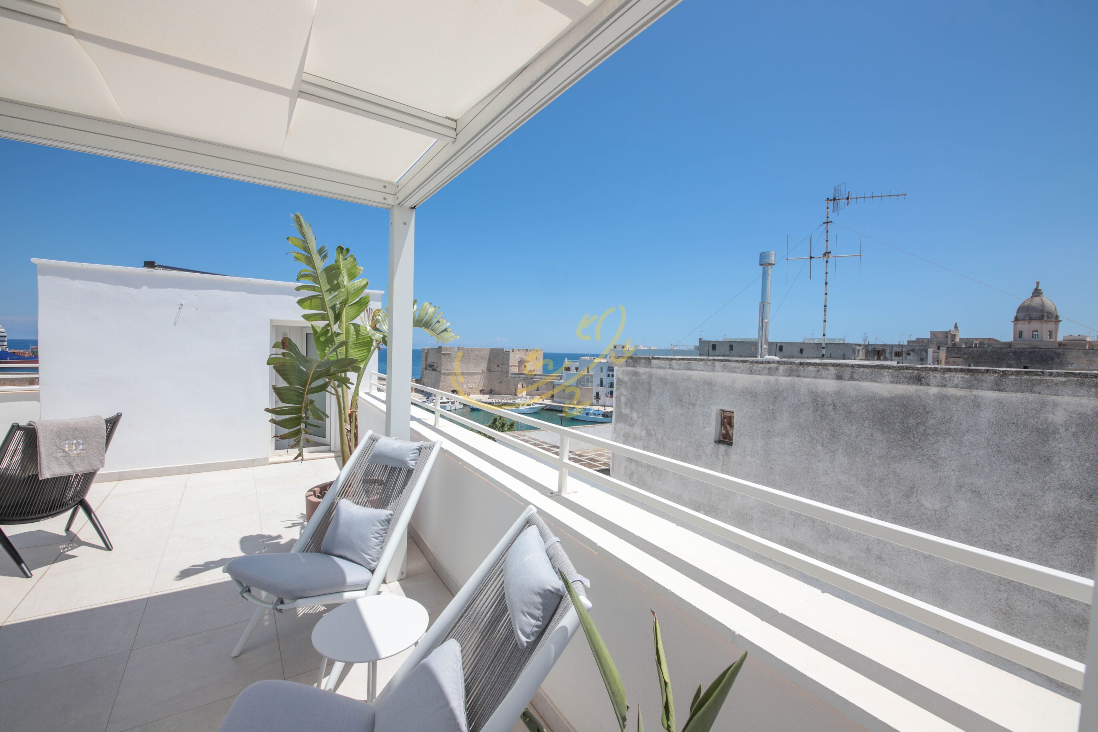 A balcony with white furniture, a plant, and a view of the sea and buildings.