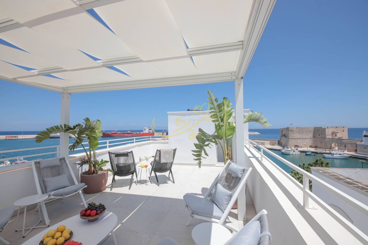 Ein Balkon mit Sitzgelegenheit, Blumentöpfen und Blick auf das Meer sowie ein historisches Gebäude.