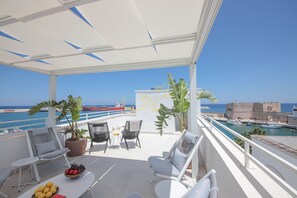 Een balkon met zitplaatsen, plantenbakken en uitzicht op zee en een historisch gebouw.