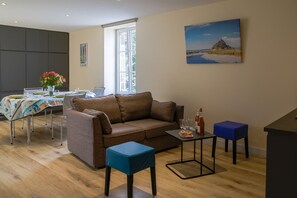 A living room with a brown sofa, a dining table with chairs, a small blue table, and a painting on the wall. - Le Mélorien by Archipel Evasion (Saint-Méloir-des-Ondes)