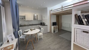 A modern kitchen with white cabinets, a dining table with chairs, a TV, and a bookshelf.