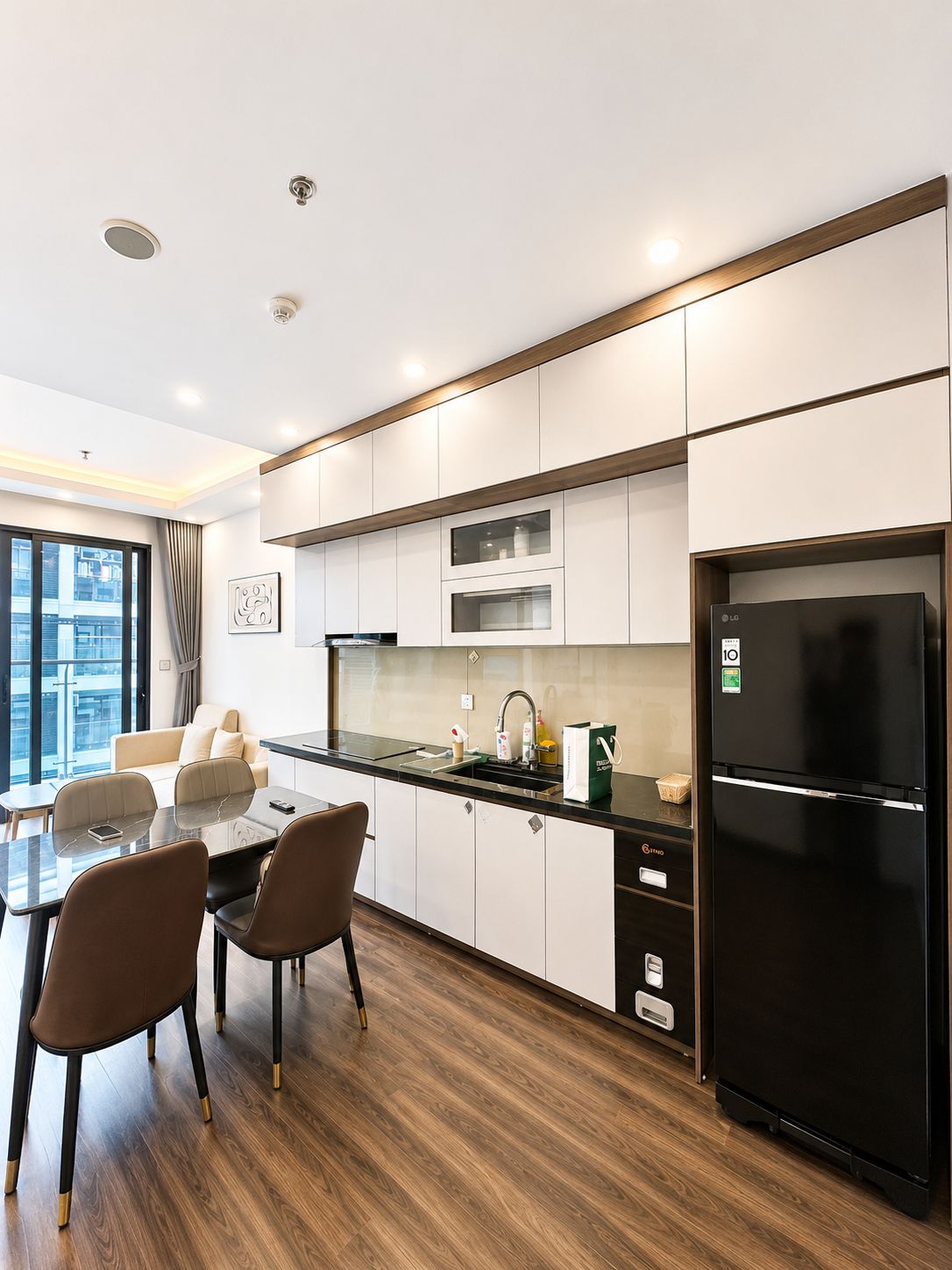 Una cocina moderna con un refrigerador negro, gabinetes blancos y un comedor con dos sillas y una mesa.