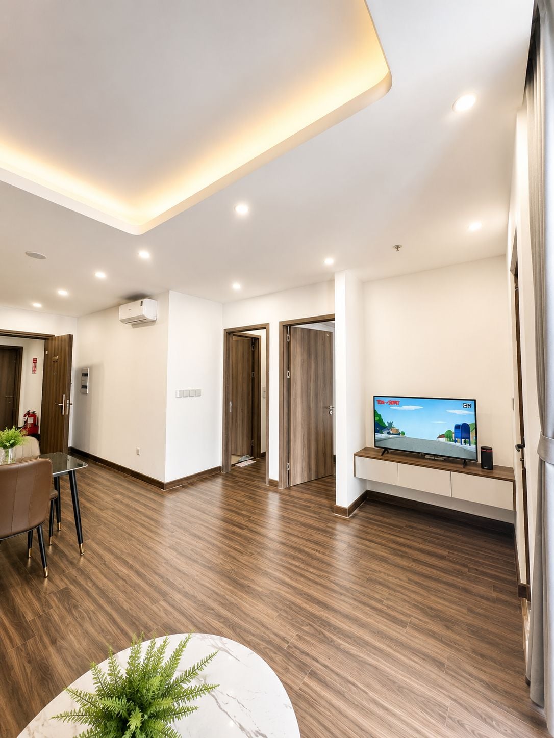 Una sala de estar moderna con un televisor de pantalla plana, un comedor con mesa y sillas, y un pasillo con puertas de madera.
