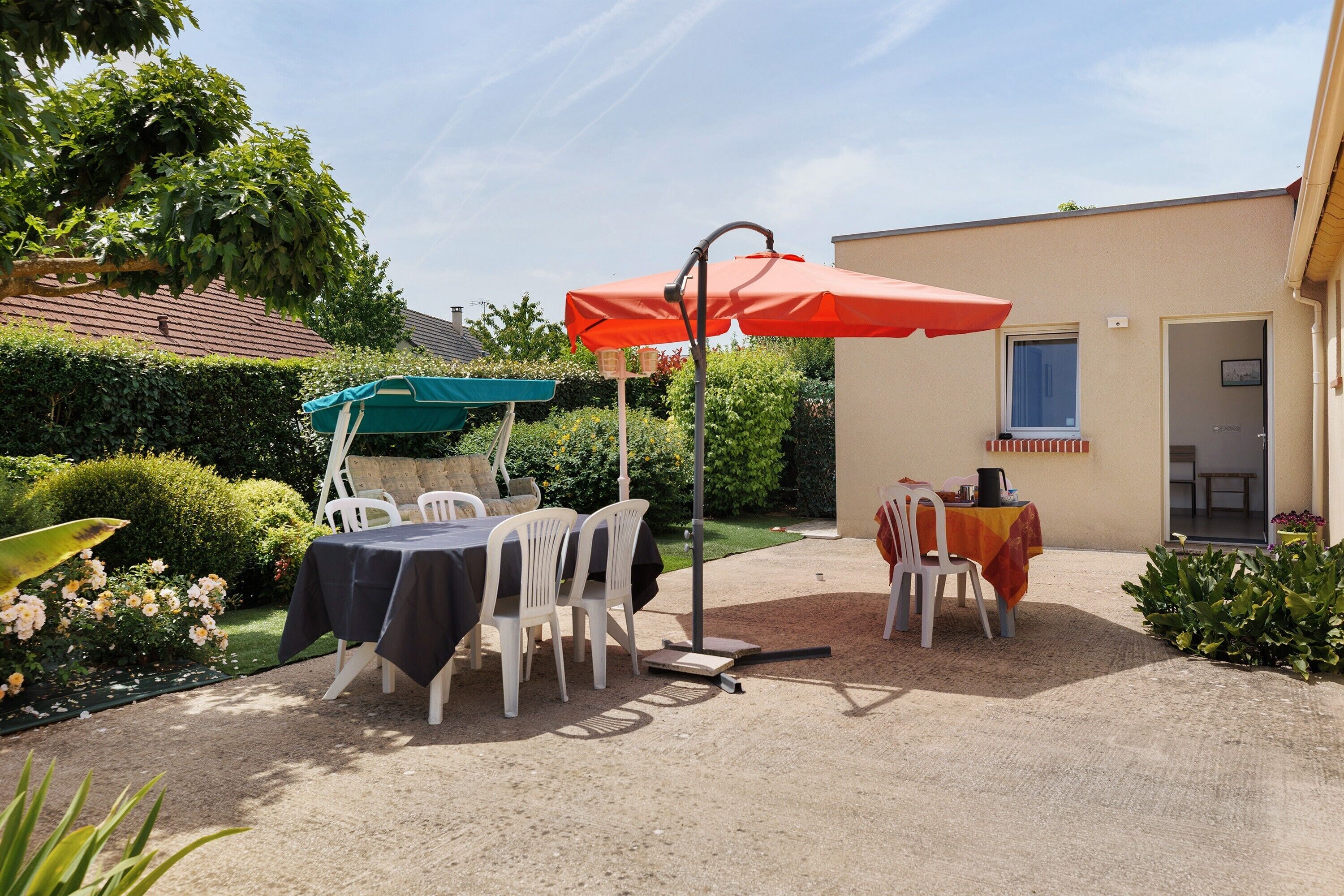 An outdoor patio with two tables, one with a red umbrella and another with a green umbrella, surrounded by white plastic chairs.