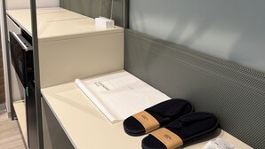 Una cama litera con un colchón blanco, una mesita con una toalla blanca y un par de pantuflas negras, y un estante de pared con una caja blanca.