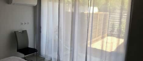 Une chambre à coucher comprenant un lit, une chaise, une fenêtre avec des rideaux légers et un appareil de climatisation.