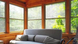 Et interiør af en træhytte med en grå sofa, store vinduer og udsigt til grøn natur.
