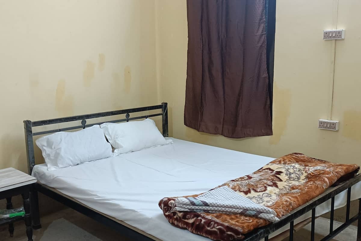 A single bed with a white mattress and a patterned blanket, a nightstand with a lamp, and a wall socket.