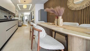 A modern kitchen with a long countertop, wooden chairs, and a dining table with a vase of dried flowers. - Beautiful 2BR seafront home with WIFI & comfy beds (Birżebbuġa)
