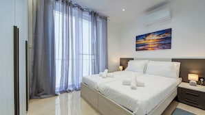 A modern hotel room with a large bed, a nightstand, a lamp, and a painting of a sunset. - Beautiful 2BR seafront home with WIFI & comfy beds (Birżebbuġa)
