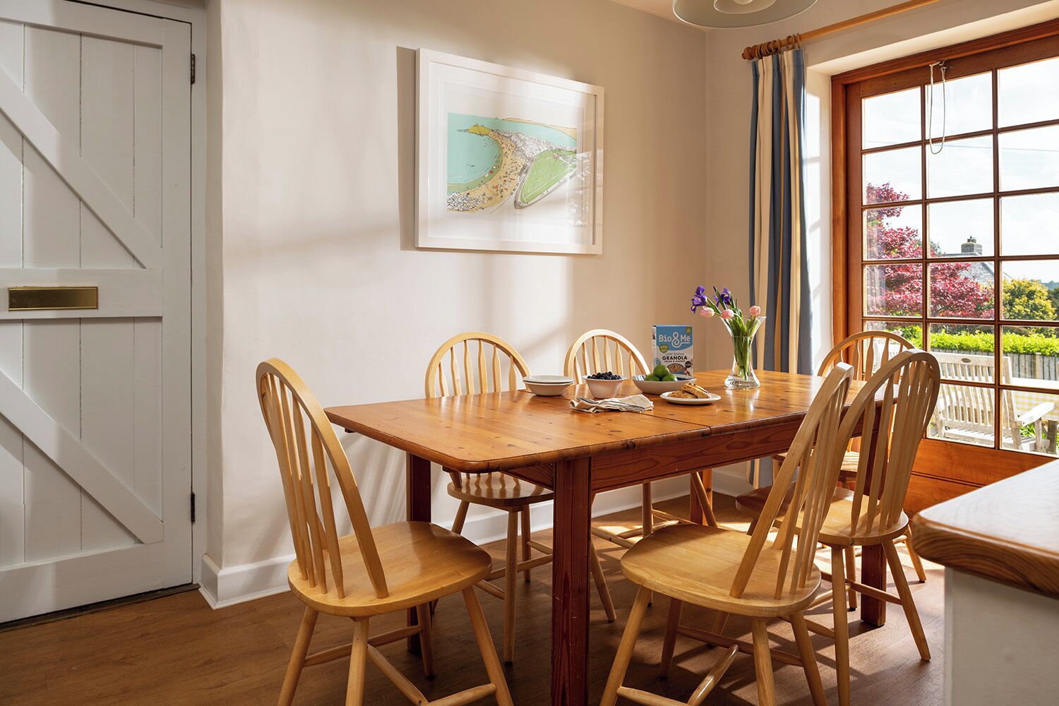 A dining area with a wooden table and chairs, a framed map on the wall, and a window with a view of greenery.
