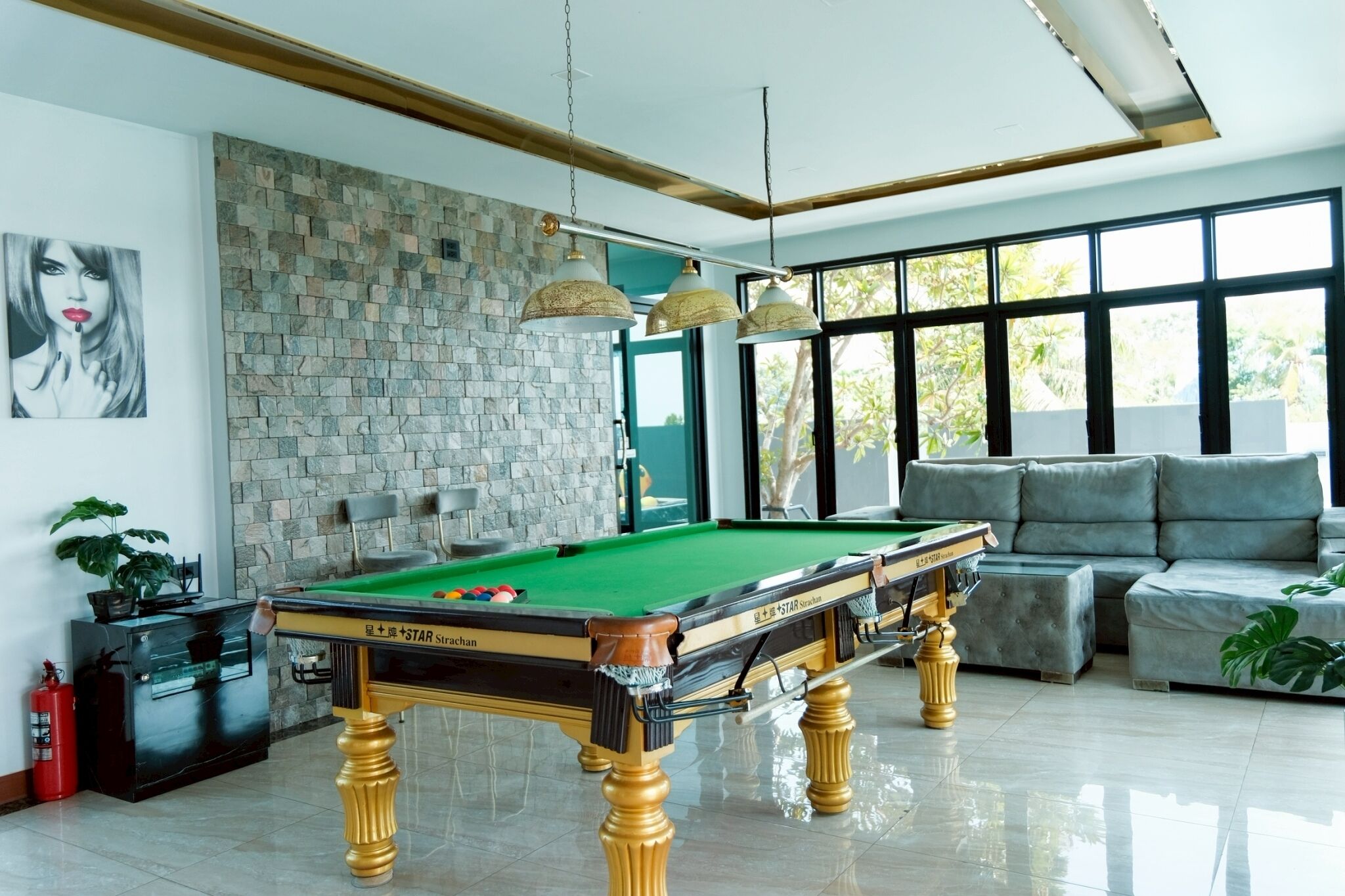 A pool table in a well-lit room with a stone wall, a sofa, and a large window.