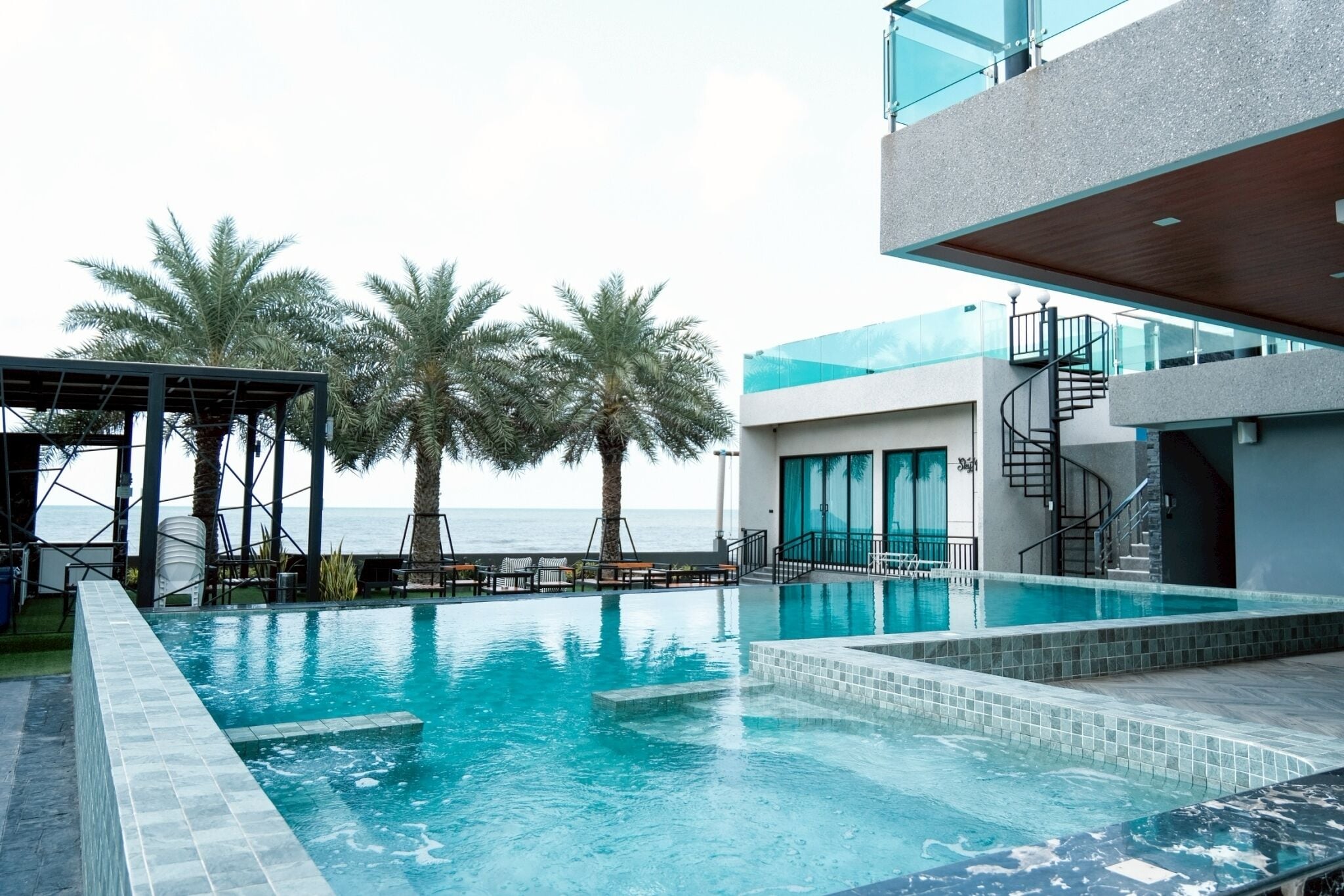 A swimming pool with a view of the sea, palm trees, and a modern building with a staircase.