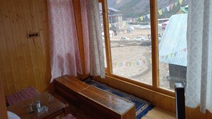 A wooden room with a bench, a table with items, and a large window with a view of buildings and mountains. - Kaas Home Stay (Chitkul)