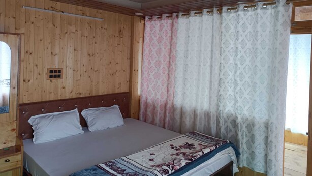 A bedroom with a wooden ceiling, a bed with white bedding, a patterned throw, and a window with a curtain. - Kaas Home Stay (Chitkul)