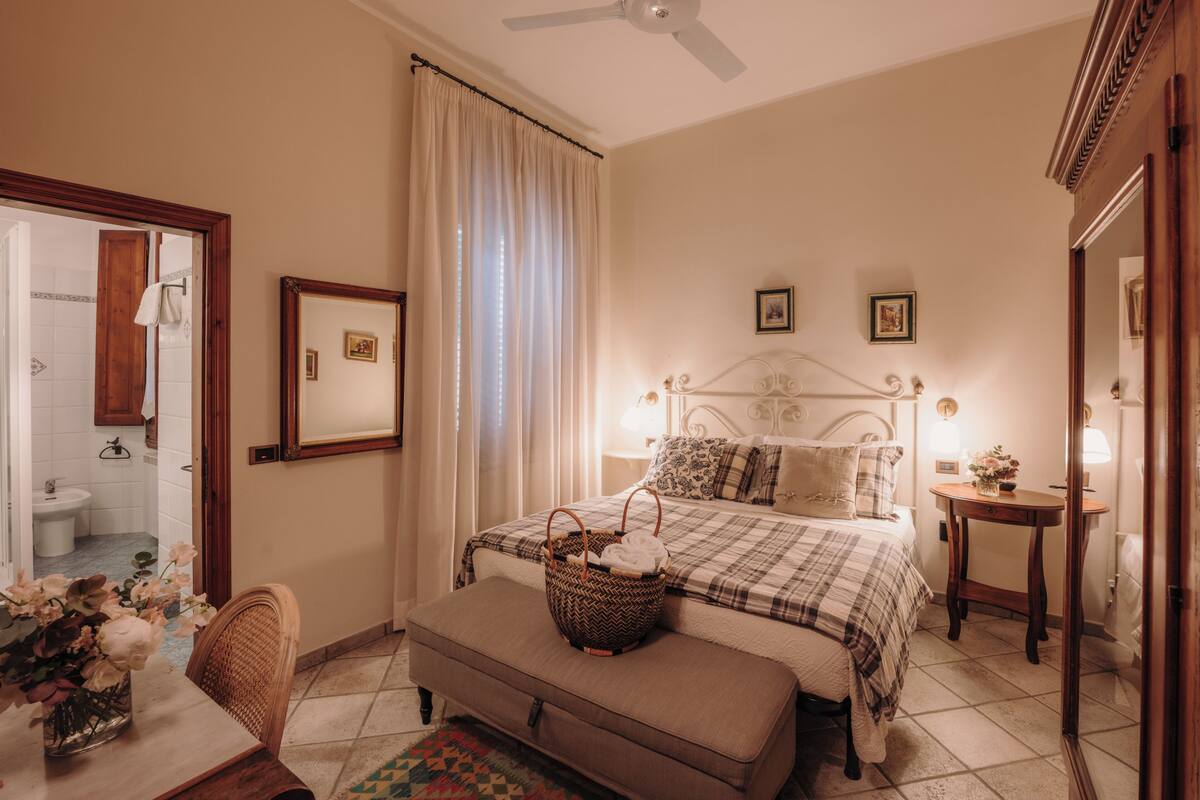 A bedroom with a bed, bedside tables, a mirror, a window with curtains, a bathroom visible through an open door, and a dining area with a table and chairs.