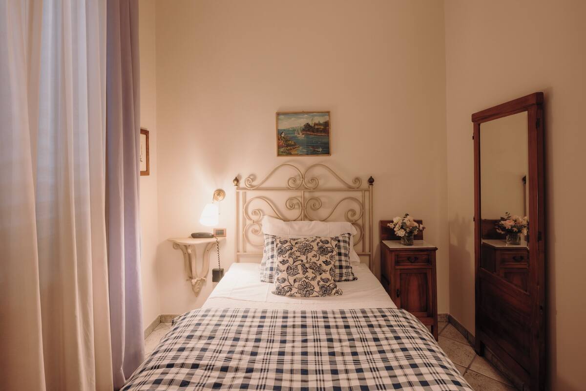 A bedroom with a bed, a nightstand, a dresser, a mirror, a painting on the wall, and a window with curtains.