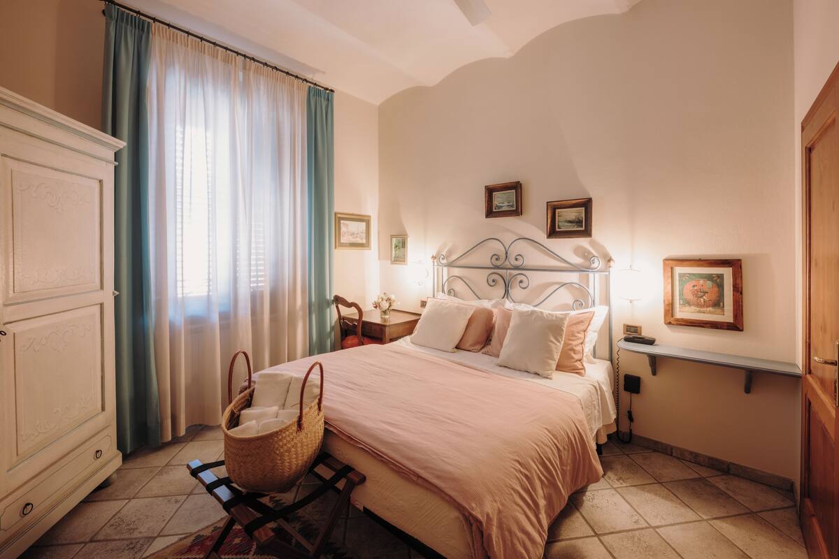A bedroom with a bed, a nightstand, a dresser, a window with curtains, and a wall-mounted shelf with pictures.